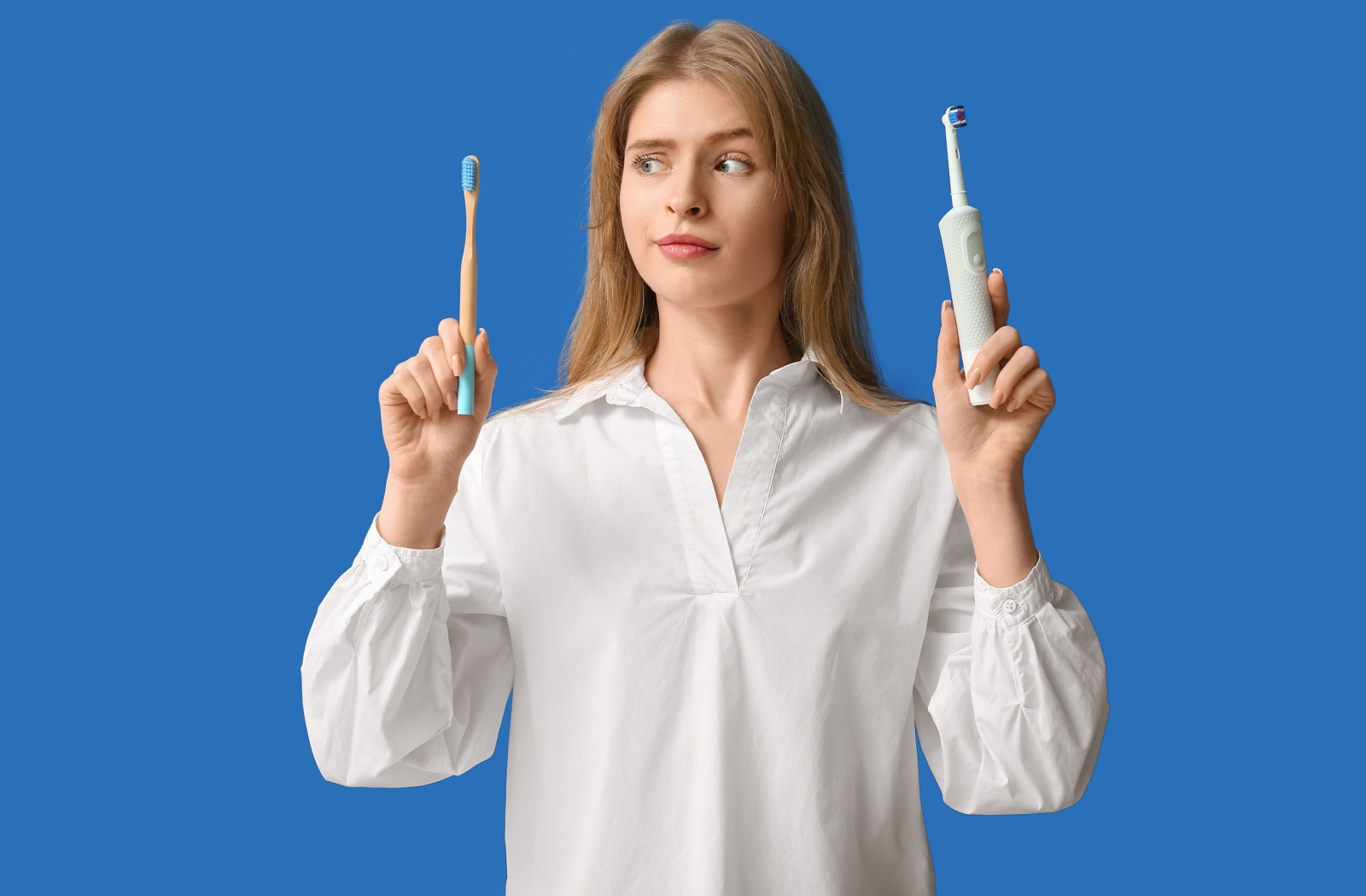 A woman with long blonde hair wearing a white blouse and holding a manual toothbrush in one hand an electric toothbrush in the other. She is raising one eyebrow.